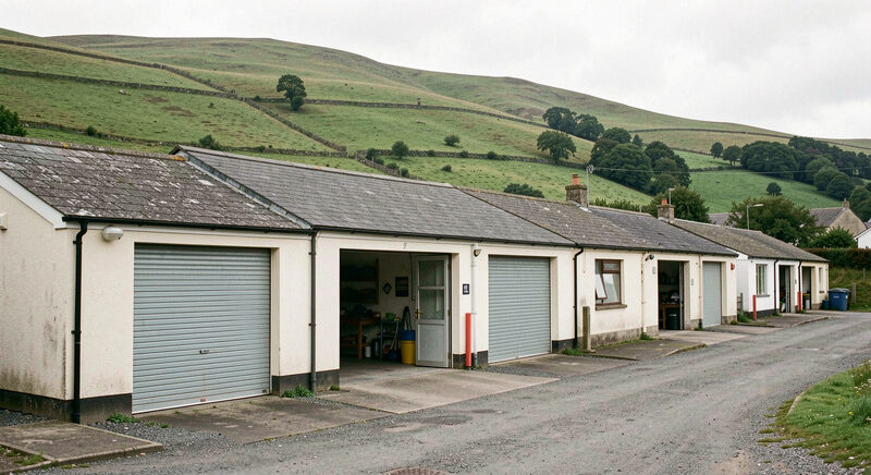 A row of small industrial units with open roller doors, green Welsh hills behind the roofs.